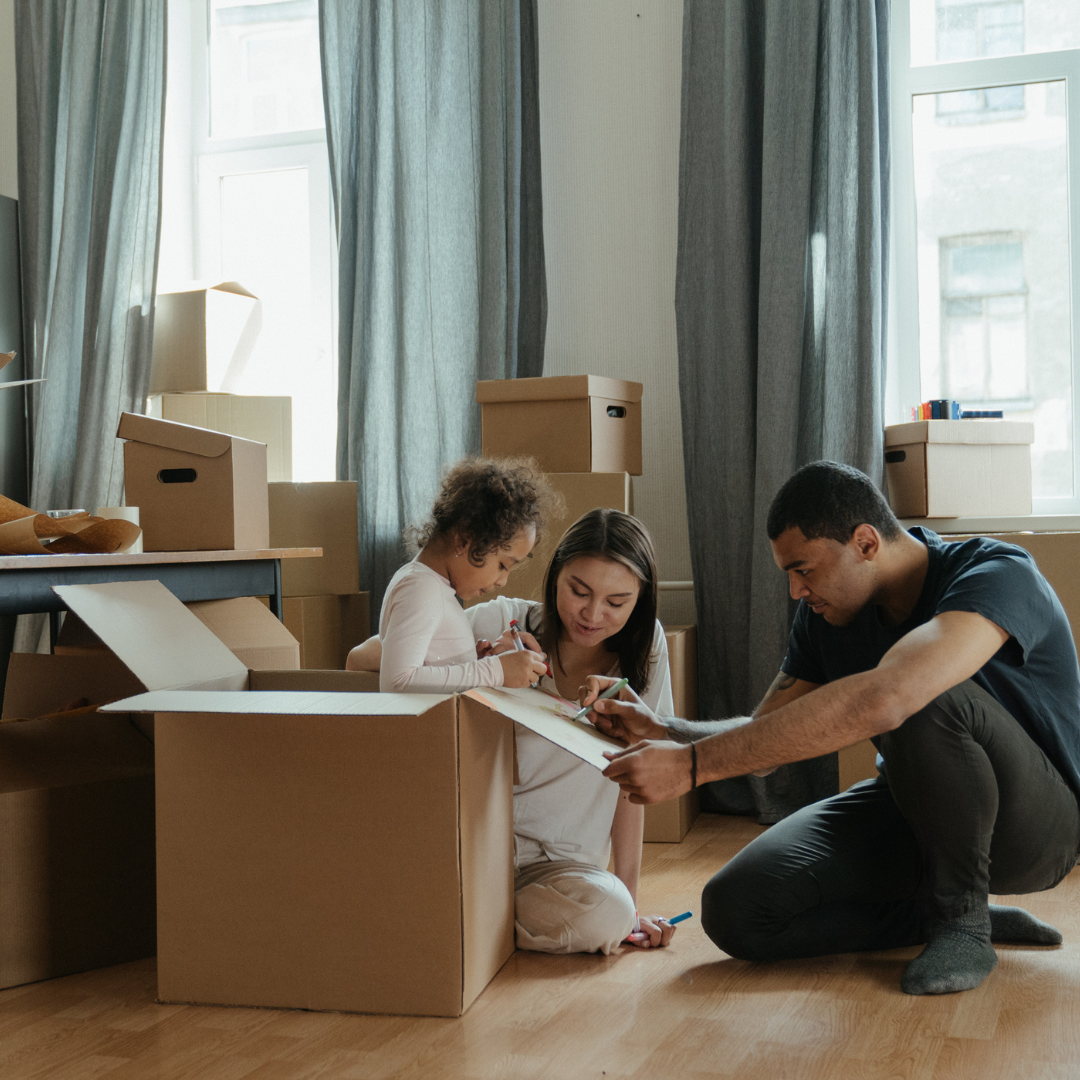 Family of 3 sitting on the floor in their new apartment smiling, while unpacking moving boxes.